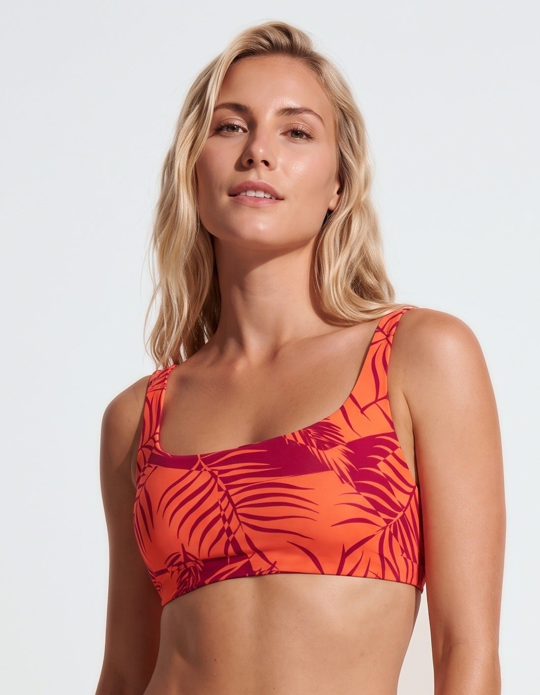 A woman modeling a two-piece bikini swimsuit with a vibrant orange and dark red tropical leaf print against a plain white studio background.