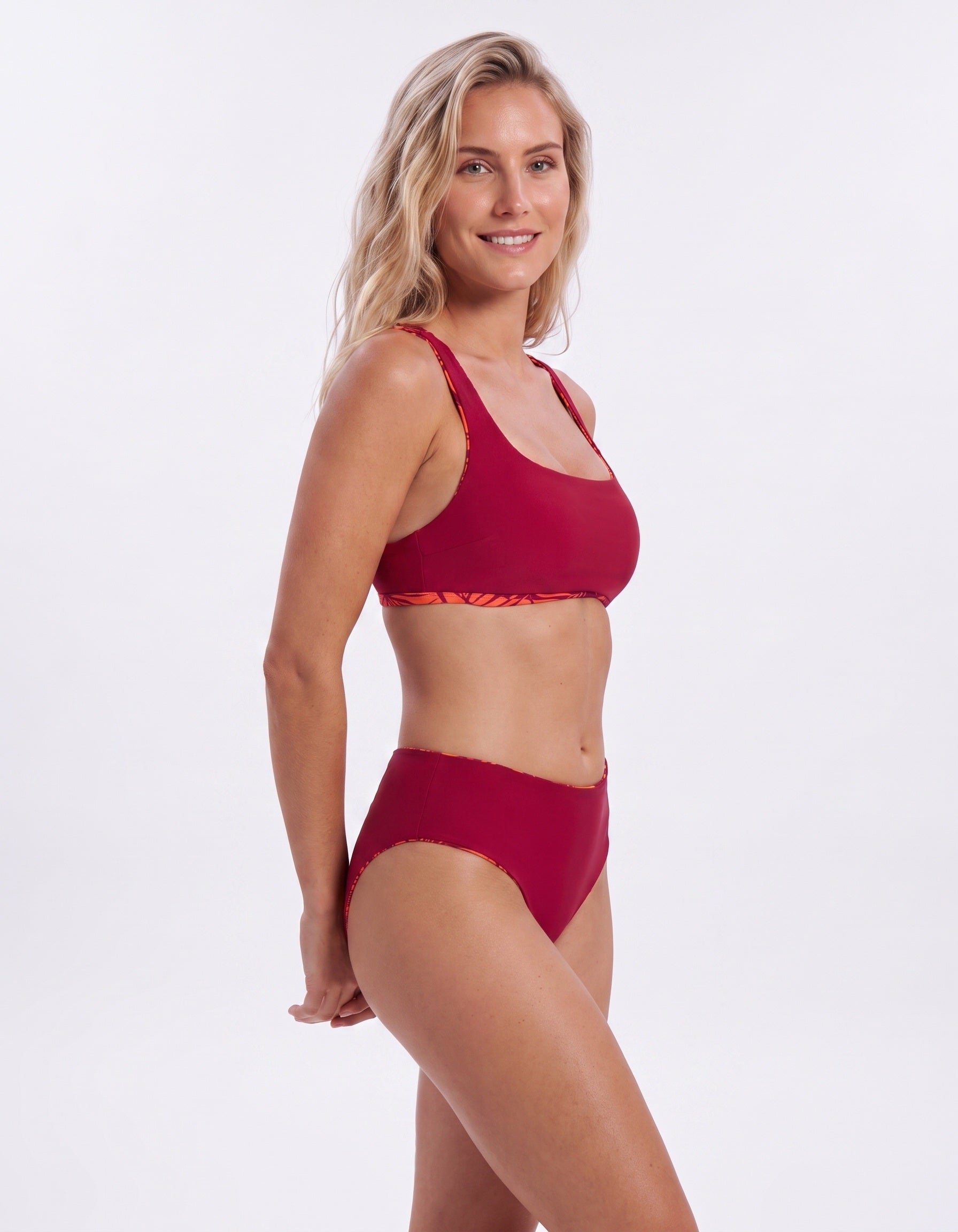 A woman modeling a two-piece bikini set in a deep red color, featuring a sports bra top with a subtle orange patterned trim and matching high-waisted bottoms, set against a plain white studio background.