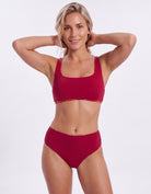 A woman modeling a matching two-piece bikini set in a deep red color with a subtle gold patterned trim along the edges, set against a plain light gray background.