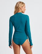 A woman shown from the back wearing a teal long-sleeve swim rash guard and matching bikini bottoms against a light gray studio background.