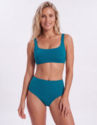 A woman modeling a two-piece swimsuit with a scoop neck bikini top and high-waisted bottoms in a solid teal color, set against a plain light gray studio background.