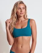 A blonde woman modeling a teal blue bikini top against a clean white studio background.