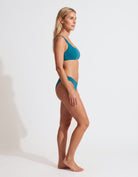 A woman in profile wearing a teal two-piece bikini swimsuit against a clean white studio background.
